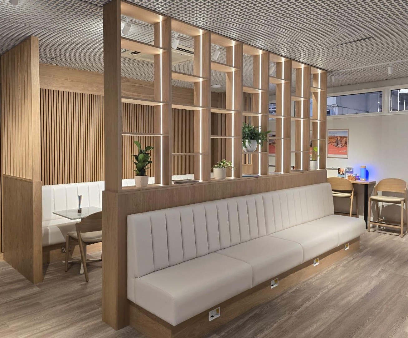 Long white fluted banquette on an oak plinth, backed by a lit oak shelving divider, with a matching booth behind set against slatted wood panelling.