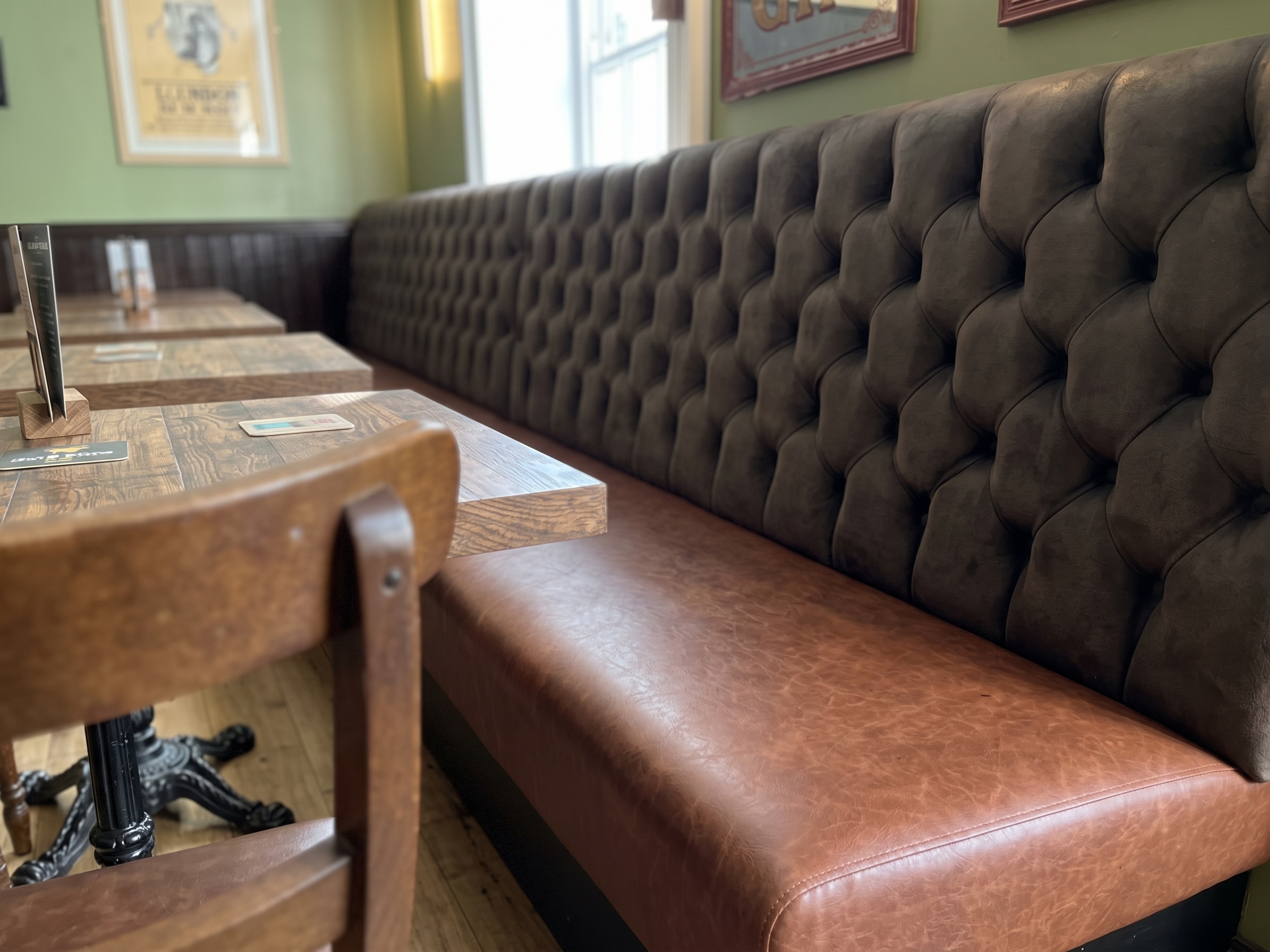 Long banquette with a deep buttoned brown velvet back and a tan leather seat, lined with rustic wood tables in a pub dining room.