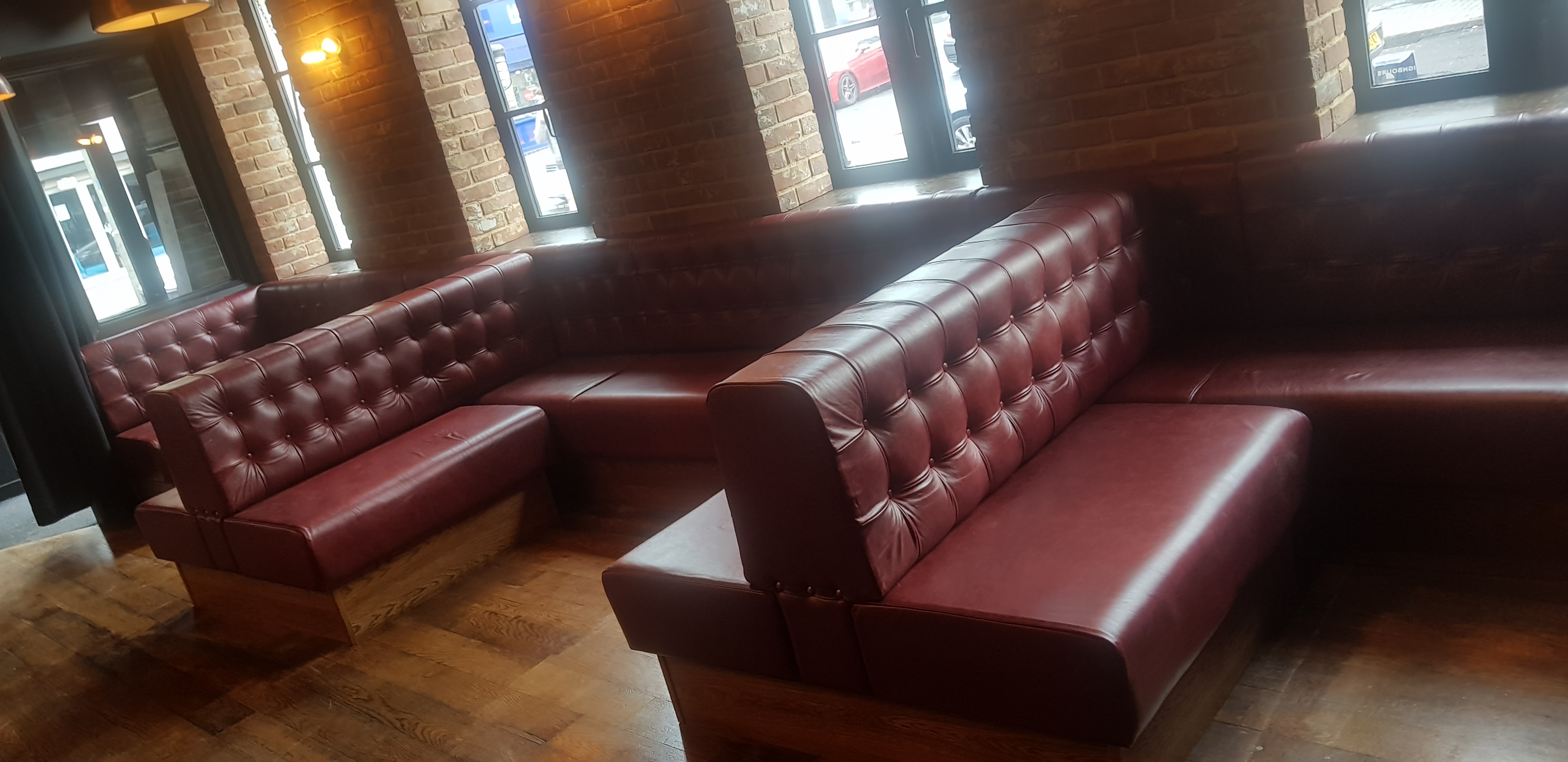 Run of deep-buttoned burgundy leather booths on oak plinths, set against the exposed brick of a city bar.